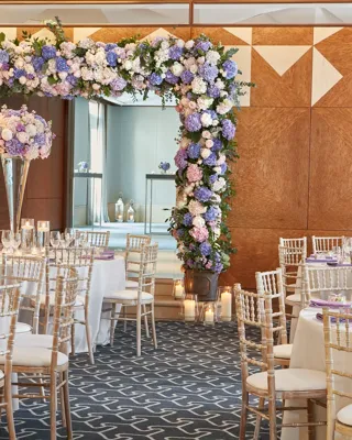Large, elegant ballroom with round tables dressed in white linens and tall gold chairs, centred on a dramatic floral arch of purple, blue, and white hydrangeas and roses, surrounded by candlelit glass votives