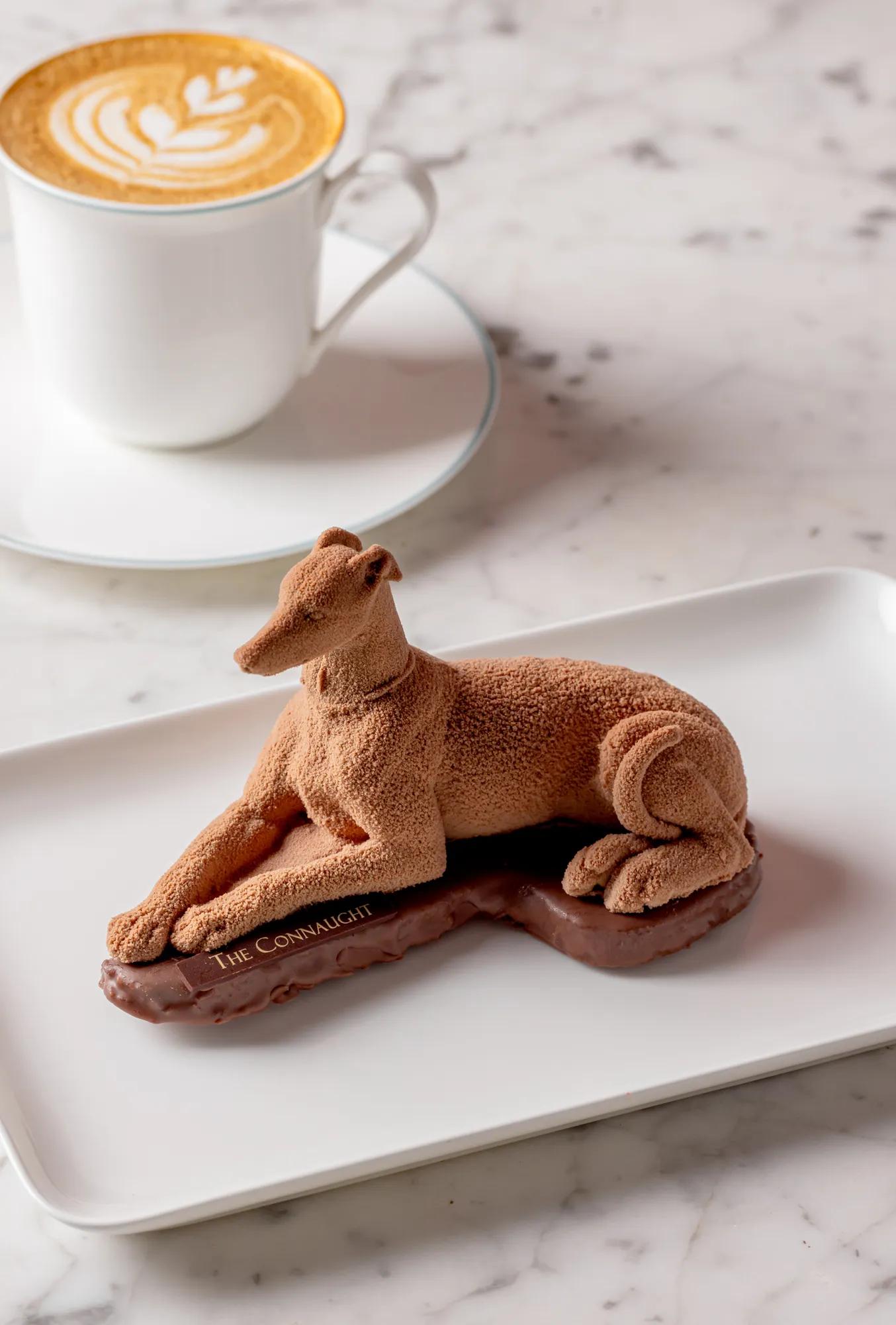 Chocolate greyhound-shaped dessert on a white plate, served alongside a cup of latte with latte art on a marble table.