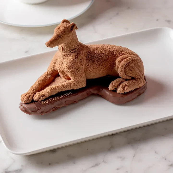 Chocolate greyhound-shaped dessert on a white plate, served alongside a cup of latte with latte art on a marble table.