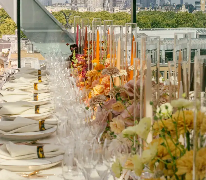 An elegant rooftop dining table with floral arrangements and glassware overlooking London’s skyline, including the Shard and London Eye.