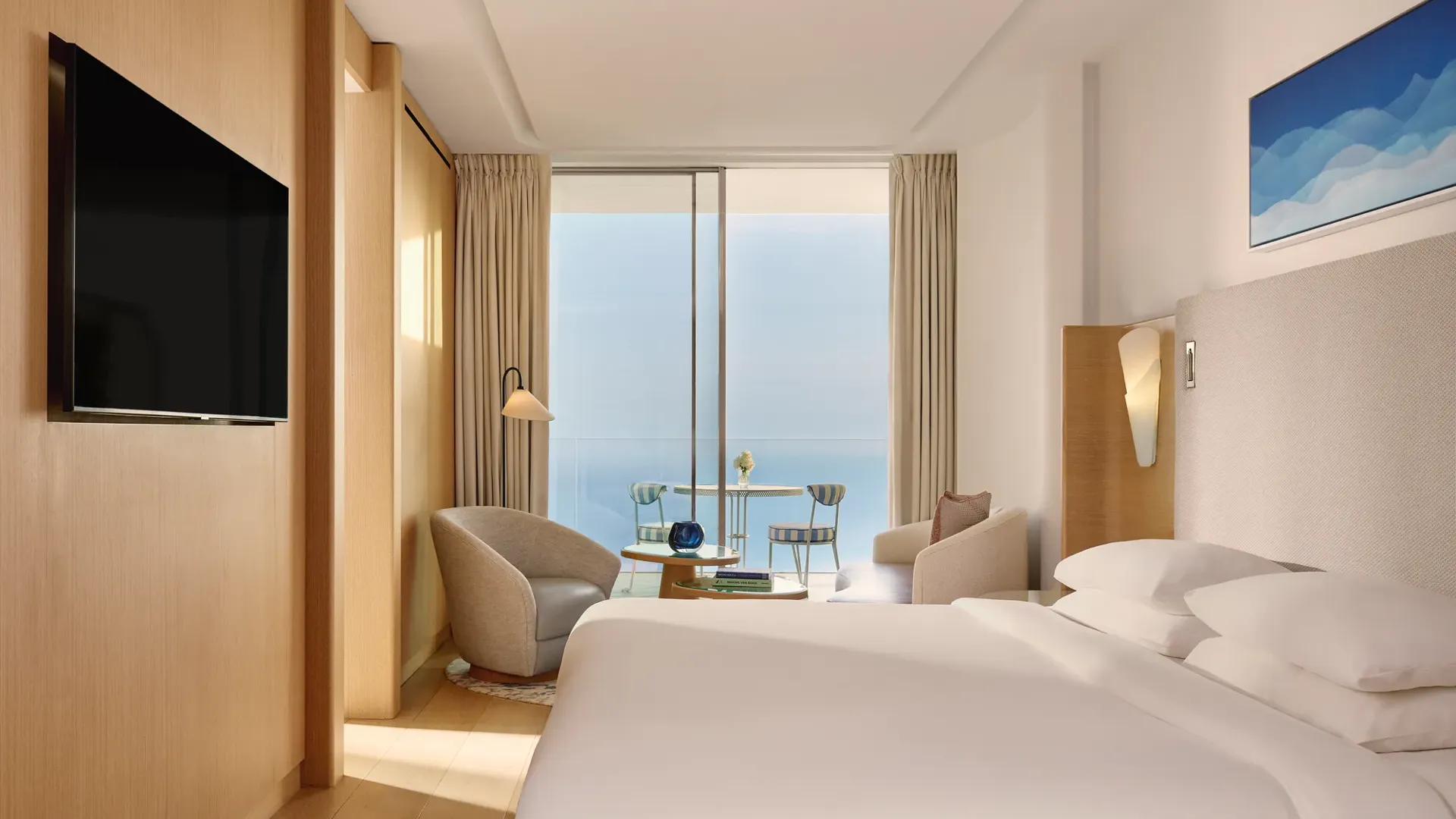 Contemporary hotel bedroom with large bed, seating area, and flat-screen TV, opening onto a terrace with sea view through sliding glass doors.