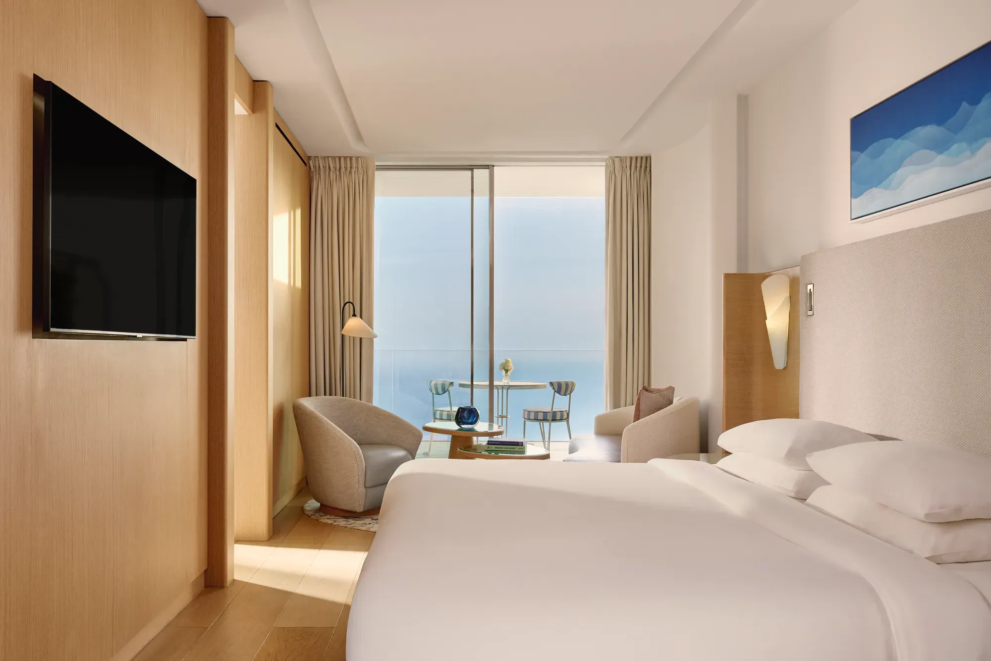 Contemporary hotel bedroom with large bed, seating area, and flat-screen TV, opening onto a terrace with sea view through sliding glass doors.