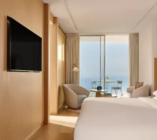 Chambre d’hôtel contemporaine avec grand lit, coin salon et télévision à écran plat, s’ouvrant sur une terrasse avec vue sur la mer par portes vitrées coulissantes.