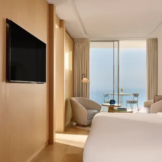 Contemporary hotel bedroom with large bed, seating area, and flat-screen TV, opening onto a terrace with sea view through sliding glass doors.