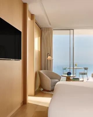 Contemporary hotel bedroom with large bed, seating area, and flat-screen TV, opening onto a terrace with sea view through sliding glass doors.