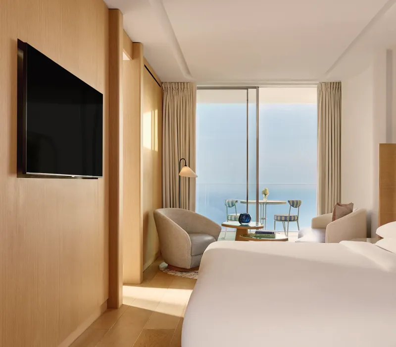 Chambre d’hôtel contemporaine avec grand lit, coin salon et télévision à écran plat, s’ouvrant sur une terrasse avec vue sur la mer par portes vitrées coulissantes.