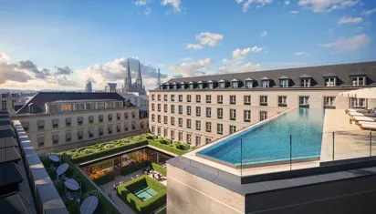 Rooftop infinity pool overlooking Paris skyline, with historic buildings, cathedral spires, and landscaped courtyard below under blue sky.