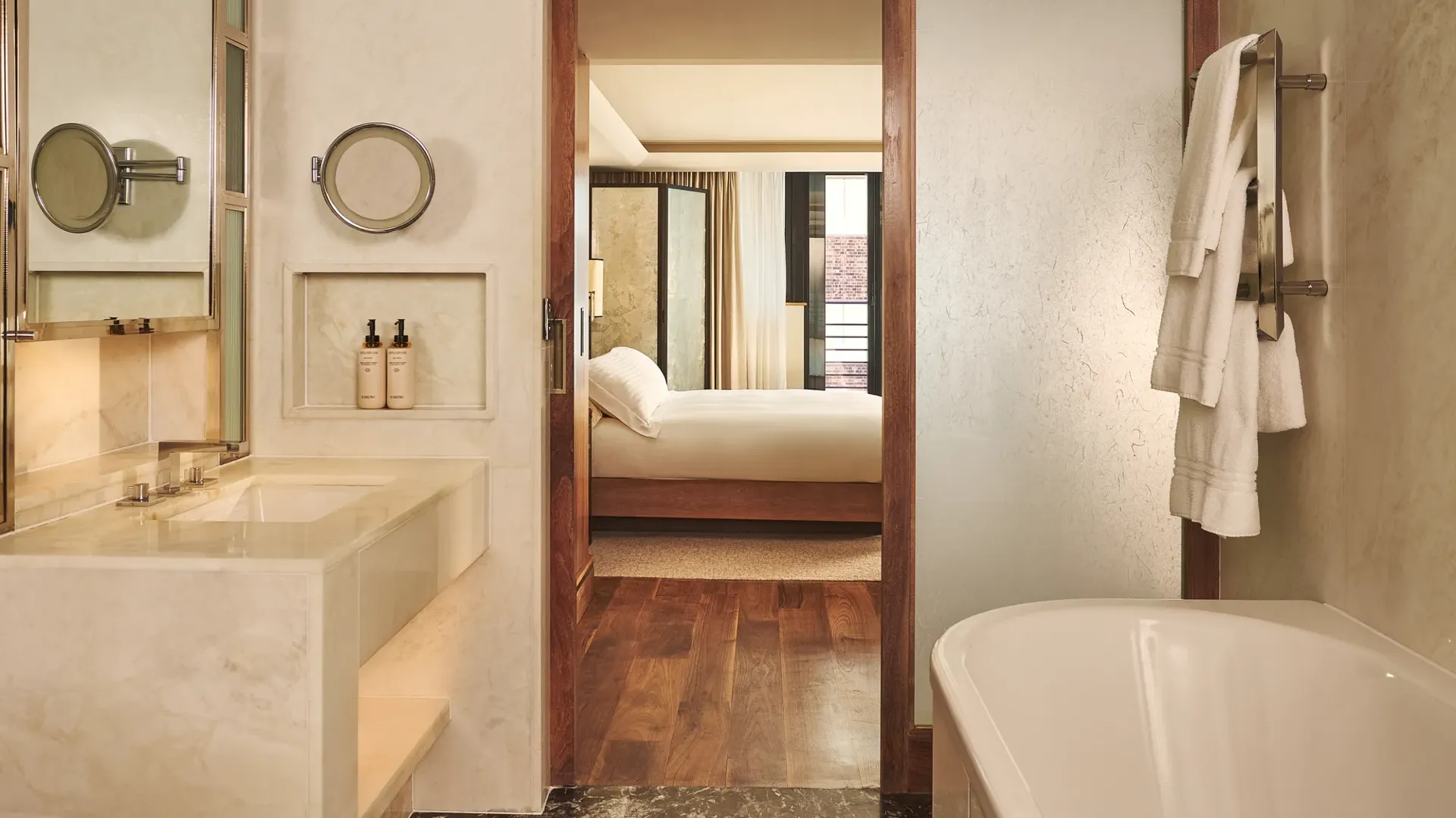 Marble bathroom with bathtub and vanity, opening through a wooden-framed door to a bedroom with a neatly made bed.