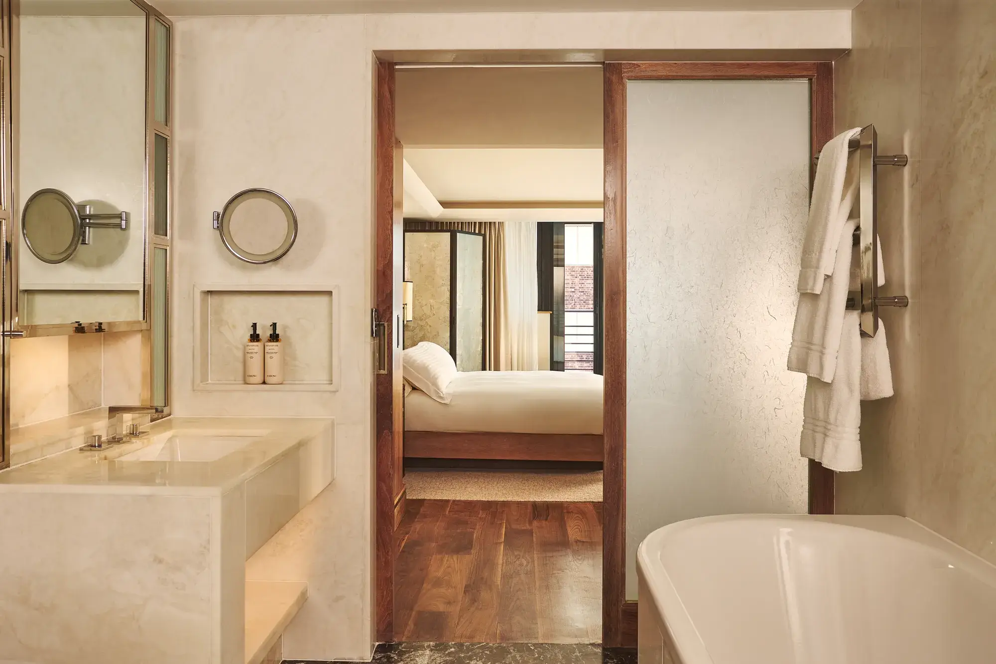 Marble bathroom with bathtub and vanity, opening through a wooden-framed door to a bedroom with a neatly made bed.