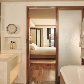 Marble bathroom with bathtub and vanity, opening through a wooden-framed door to a bedroom with a neatly made bed.