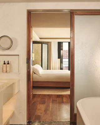 Marble bathroom with bathtub and vanity, opening through a wooden-framed door to a bedroom with a neatly made bed.