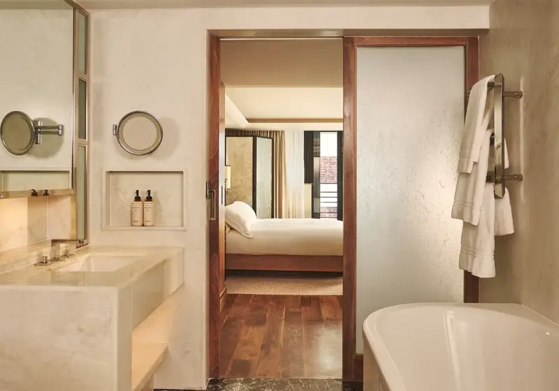 Marble bathroom with bathtub and vanity, opening through a wooden-framed door to a bedroom with a neatly made bed.