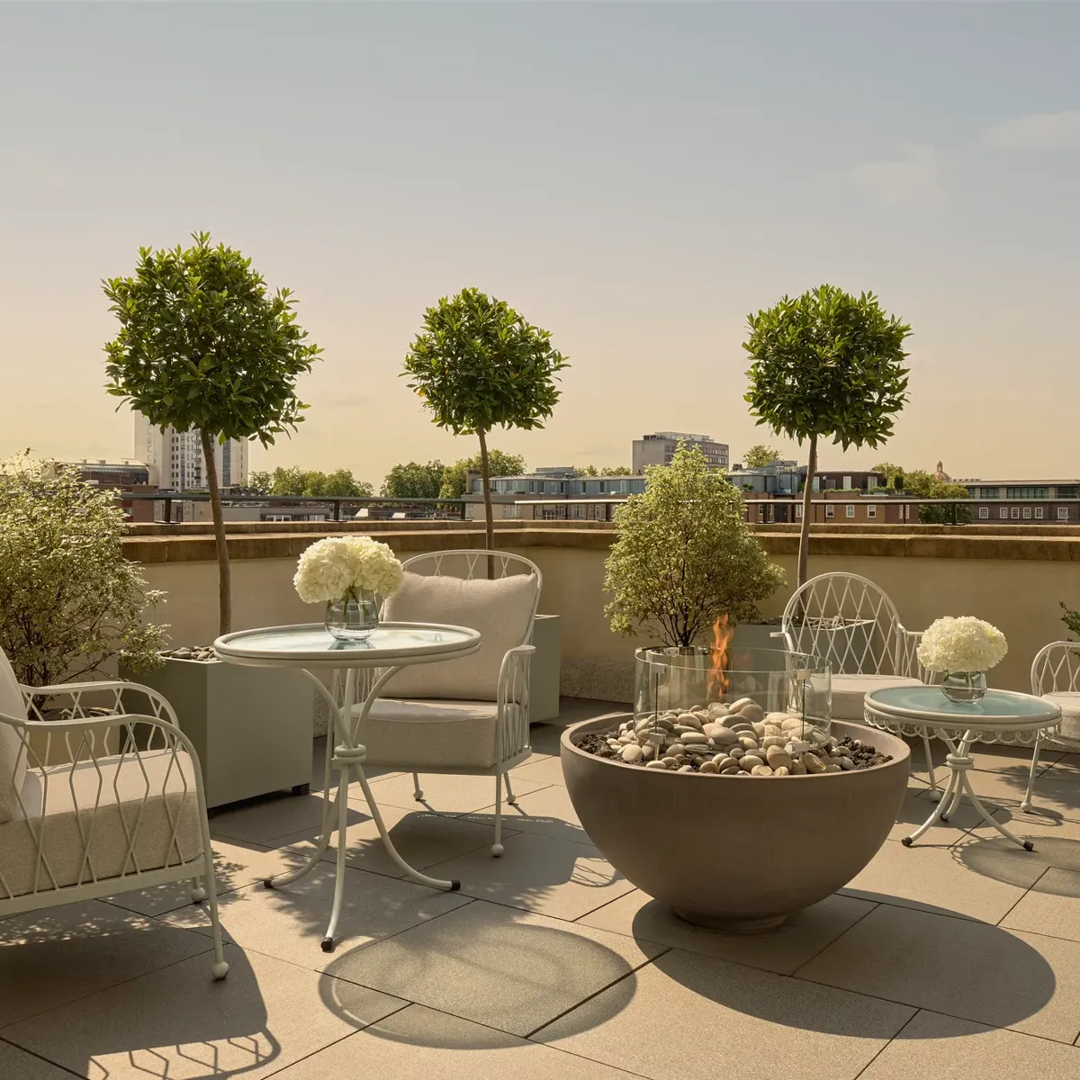 Rooftop terrace with cushioned seating, round tables, potted trees, and central fire pit overlooking city skyline.