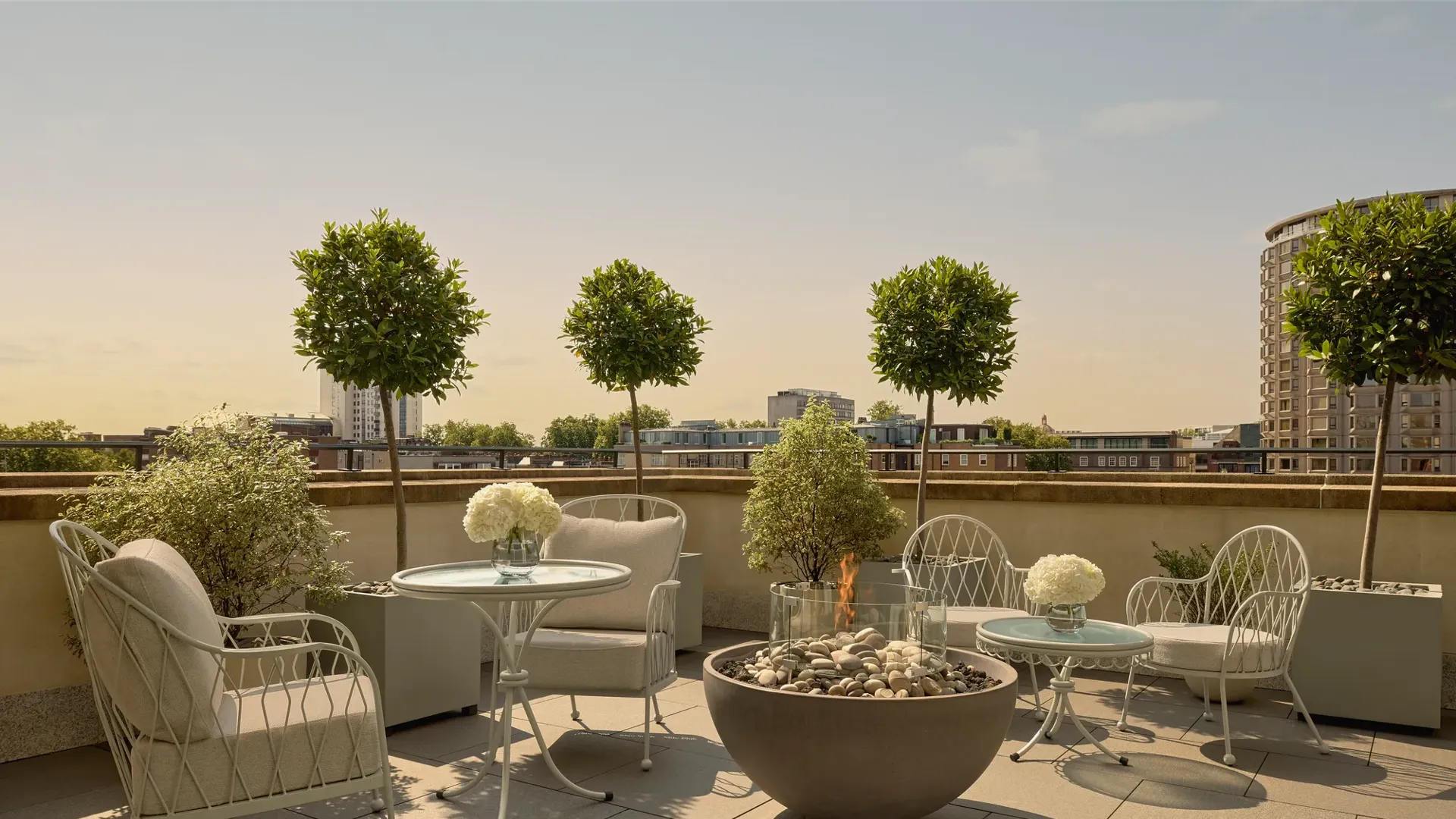 Rooftop terrace with cushioned seating, round tables, potted trees, and central fire pit overlooking city skyline.