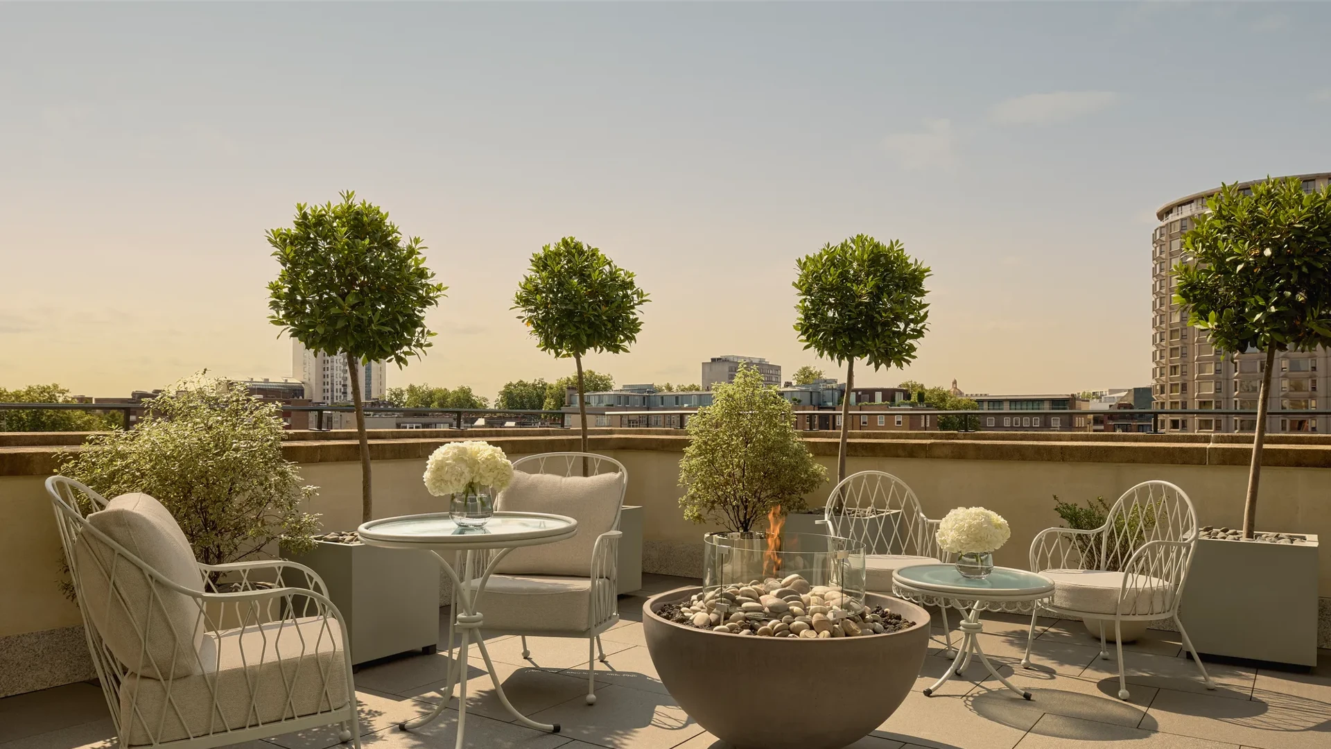 Rooftop terrace with cushioned seating, round tables, potted trees, and central fire pit overlooking city skyline.