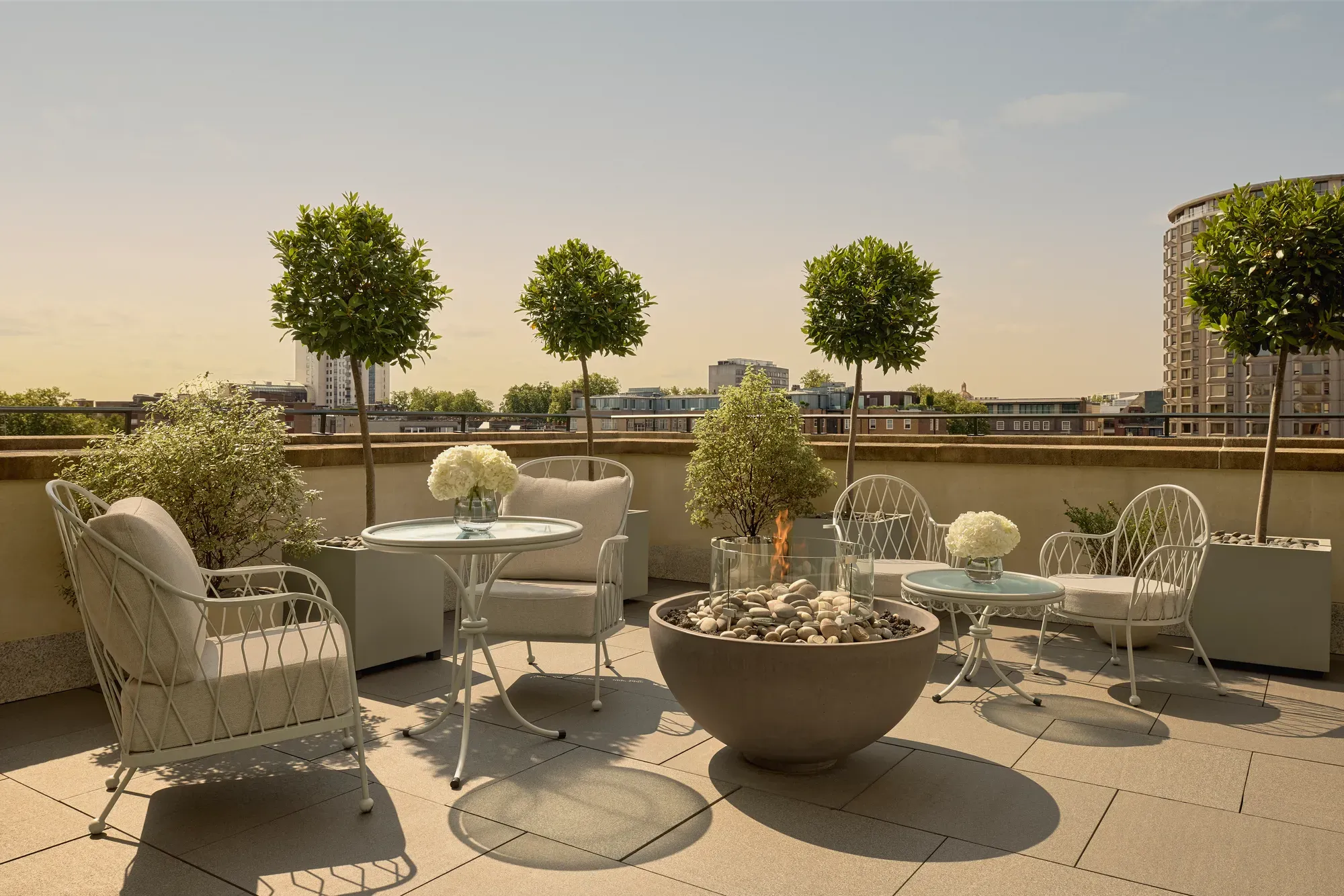 Rooftop terrace with cushioned seating, round tables, potted trees, and central fire pit overlooking city skyline.