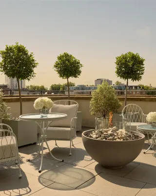 Rooftop terrace with cushioned seating, round tables, potted trees, and central fire pit overlooking city skyline.