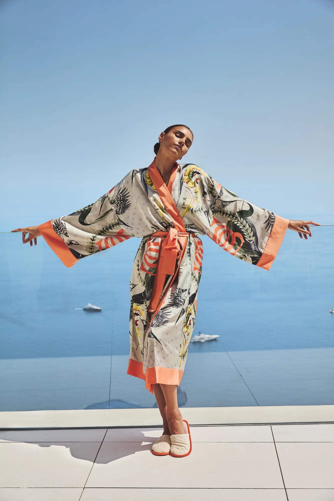 Femme en robe imprimée, bras ouverts sur une terrasse surplombant une mer bleue et paisible.