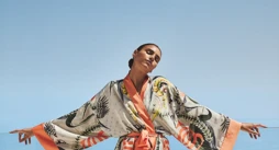 Woman in a patterned robe standing with arms open on a terrace overlooking a calm blue sea.