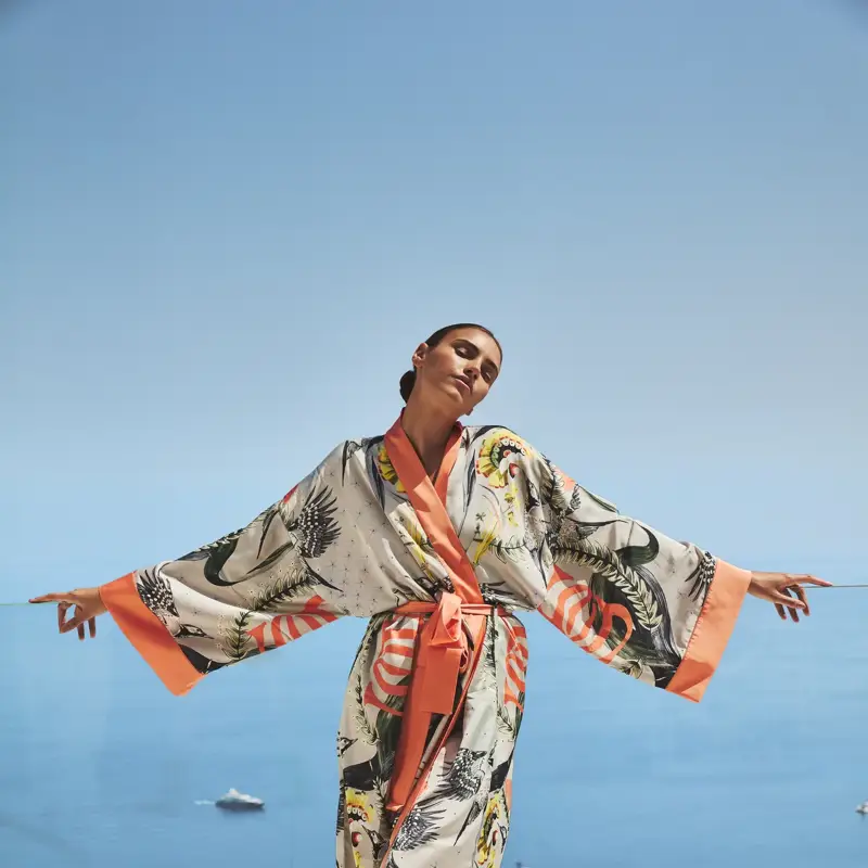 Woman in a patterned robe standing with arms open on a terrace overlooking a calm blue sea.