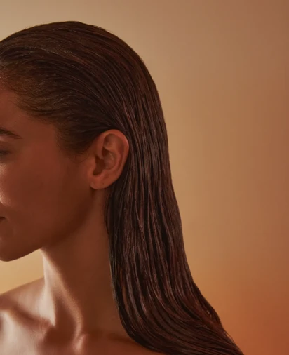 Close-up side profile of a woman with slicked-back wet hair and bare shoulders against a warm, softly lit background.