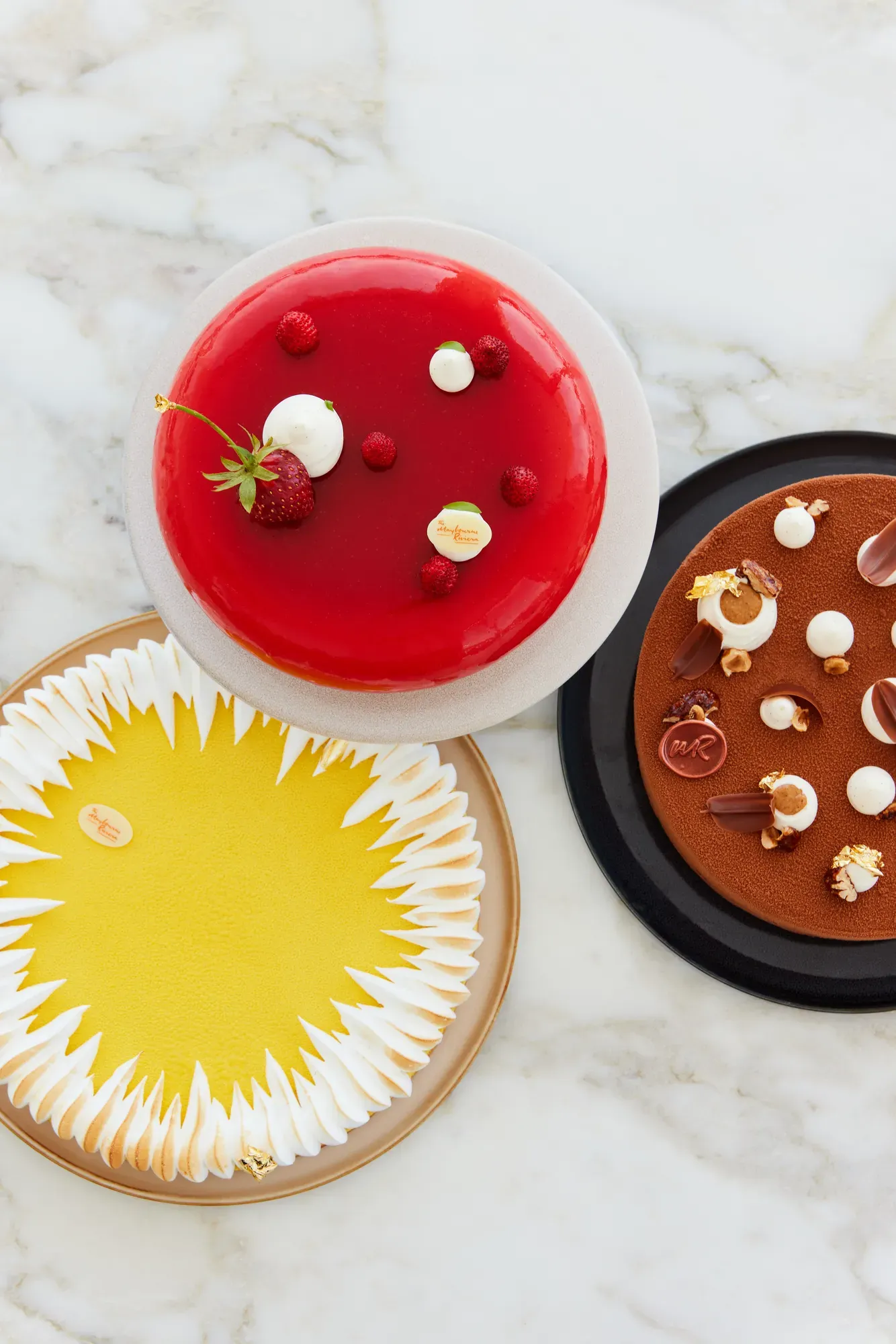 Three elegant pâtisserie cakes—red berry, yellow citrus meringue, and chocolate—arranged on a marble surface.