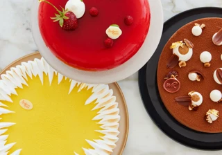 Three elegant pâtisserie cakes—red berry, yellow citrus meringue, and chocolate—arranged on a marble surface.