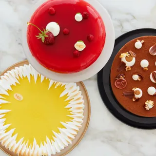 Three elegant pâtisserie cakes—red berry, yellow citrus meringue, and chocolate—arranged on a marble surface.