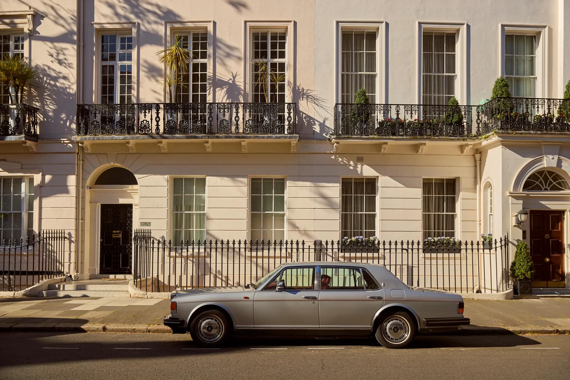 Classic silver luxury sedan parked outside elegant white Georgian townhouse with wrought-iron balconies on a sunlit residential street.
