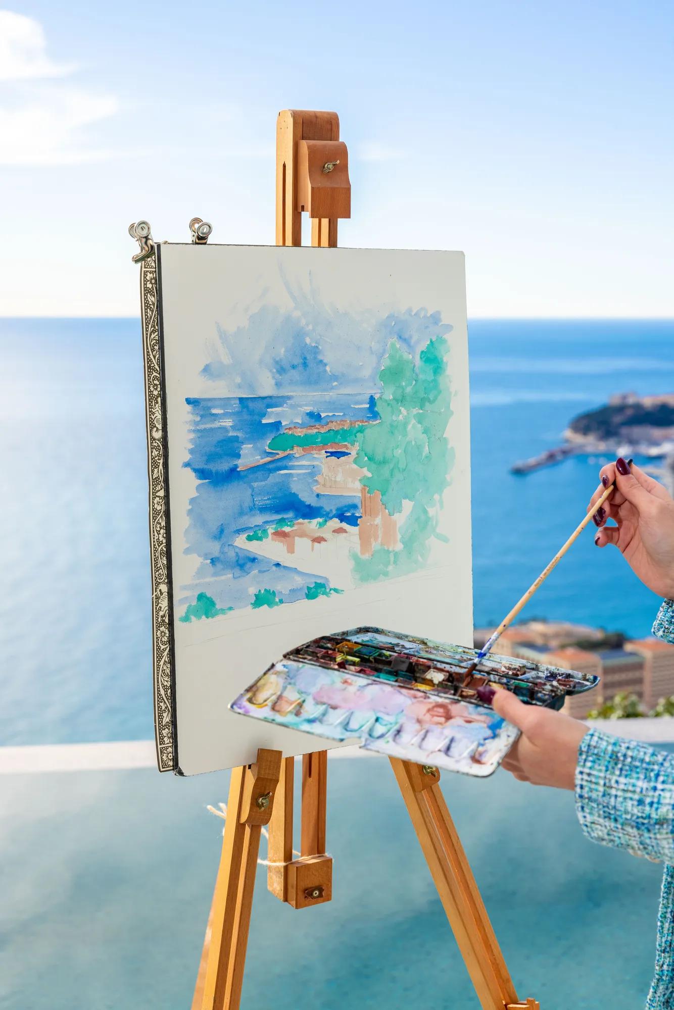 Artiste peignant un paysage marin sur chevalet, avec aquarelles et palette, dominant l’océan depuis une terrasse.