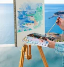 Artiste peignant un paysage marin sur chevalet, avec aquarelles et palette, dominant l’océan depuis une terrasse.