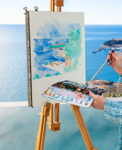 Artiste peignant un paysage marin sur chevalet, avec aquarelles et palette, dominant l’océan depuis une terrasse.