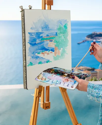 Artiste peignant un paysage marin sur chevalet, avec aquarelles et palette, dominant l’océan depuis une terrasse.