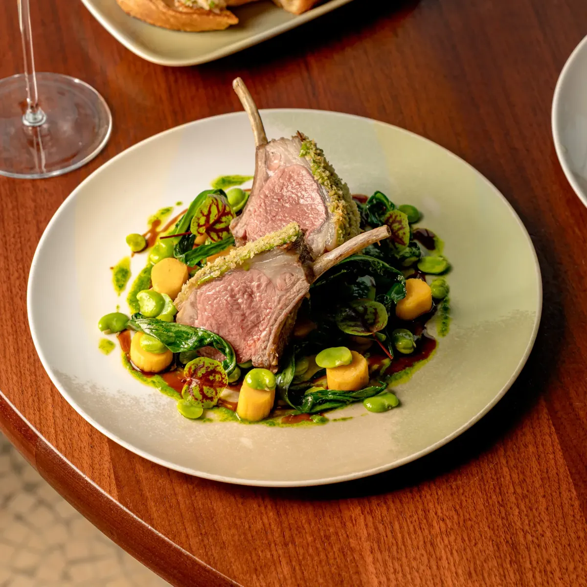 Herb-crusted lamb rack with spring vegetables, peas, and greens on a white plate with sauce, set on a wooden table