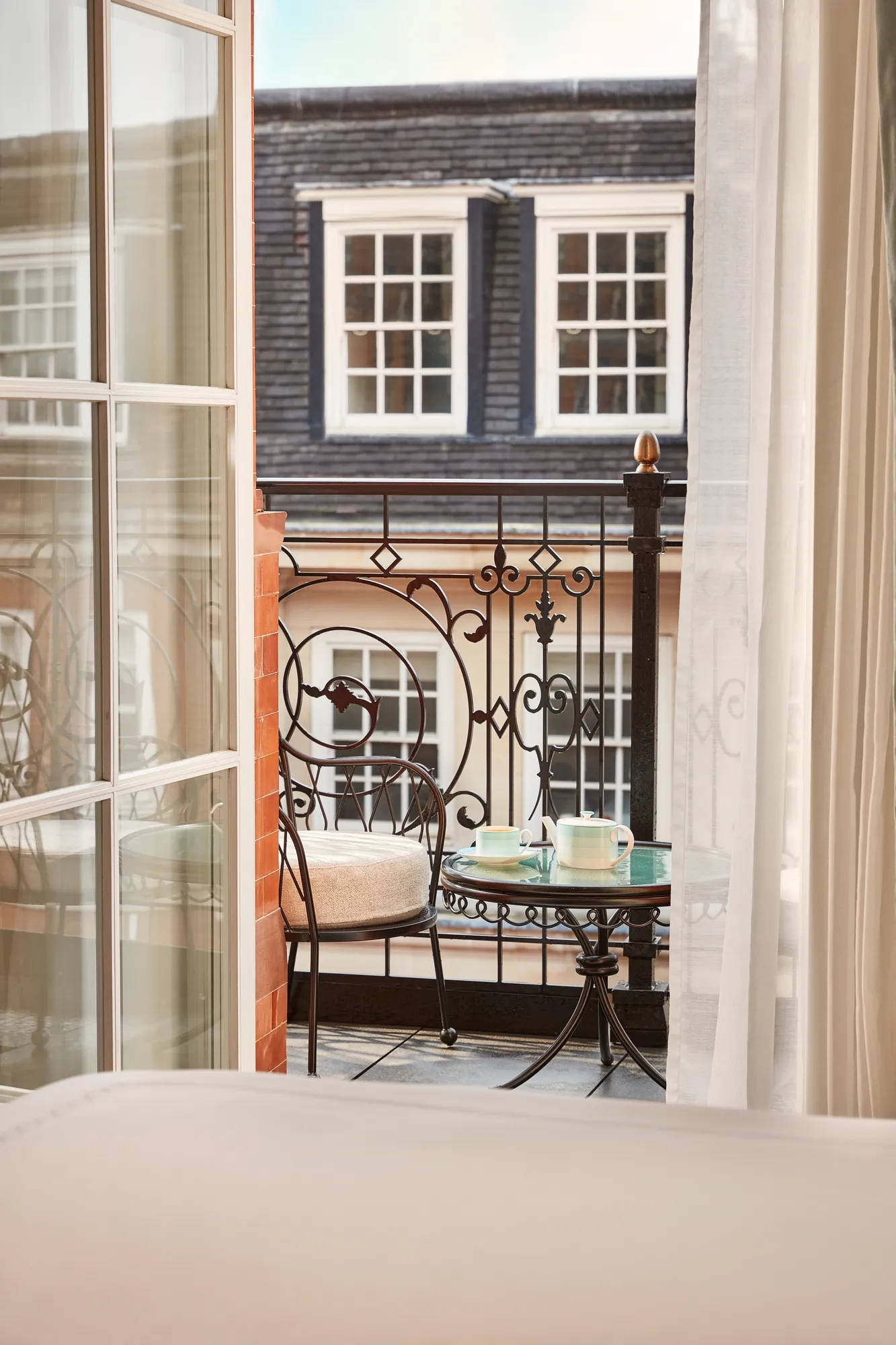 Intimate balcony with wrought-iron railing, a small round table set with teacups, and two cushioned chairs overlooking classic townhouse windows.