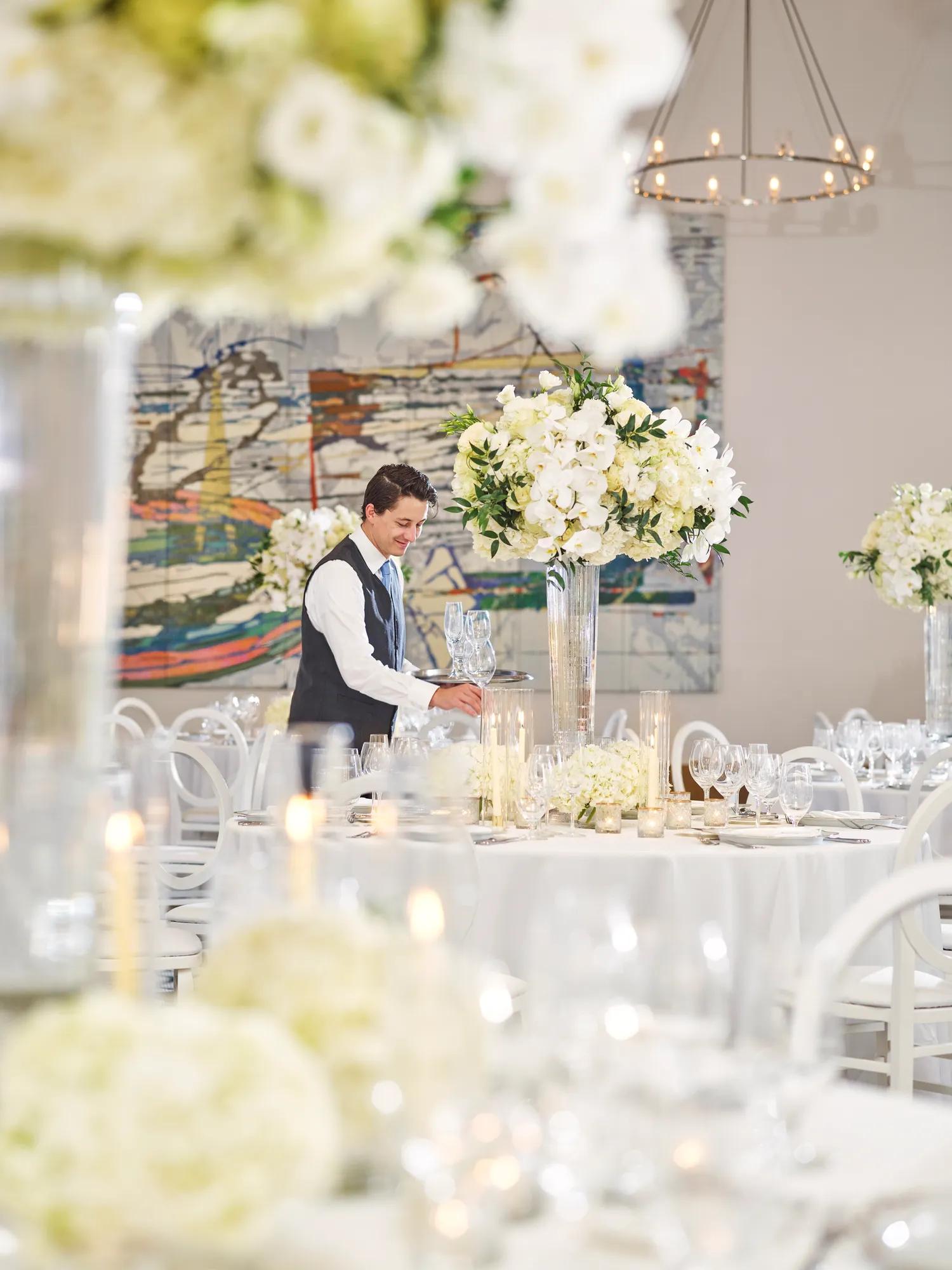 Server setting glassware among tall white floral centerpieces in a ballroom with chandeliers and an abstract mural backdrop.