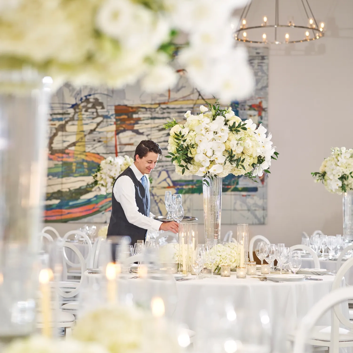 Server setting glassware among tall white floral centerpieces in a ballroom with chandeliers and an abstract mural backdrop.