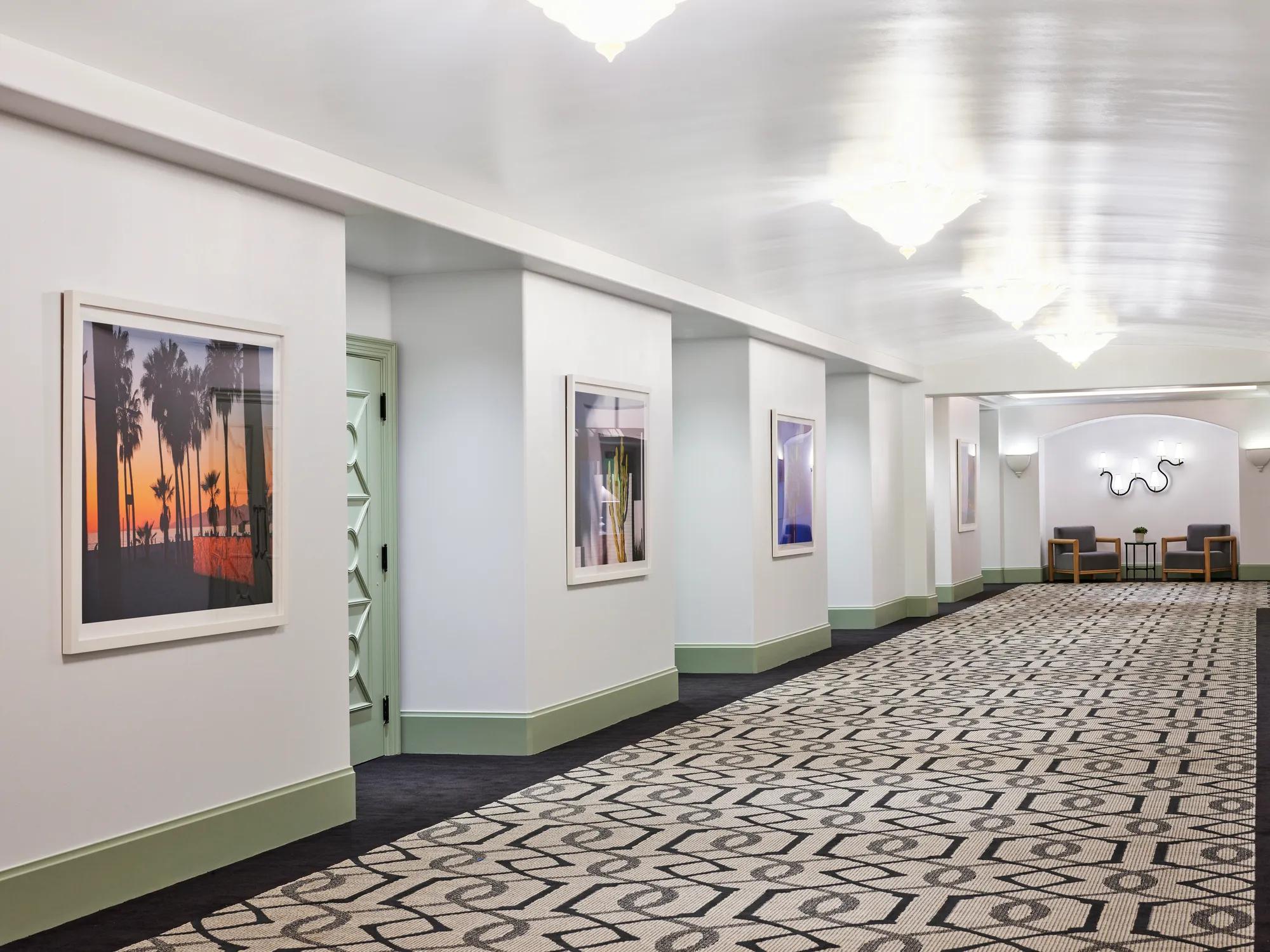 Gallery pre-function corridor with framed art, patterned carpet, mint trim, chandeliers, and small seating nooks.