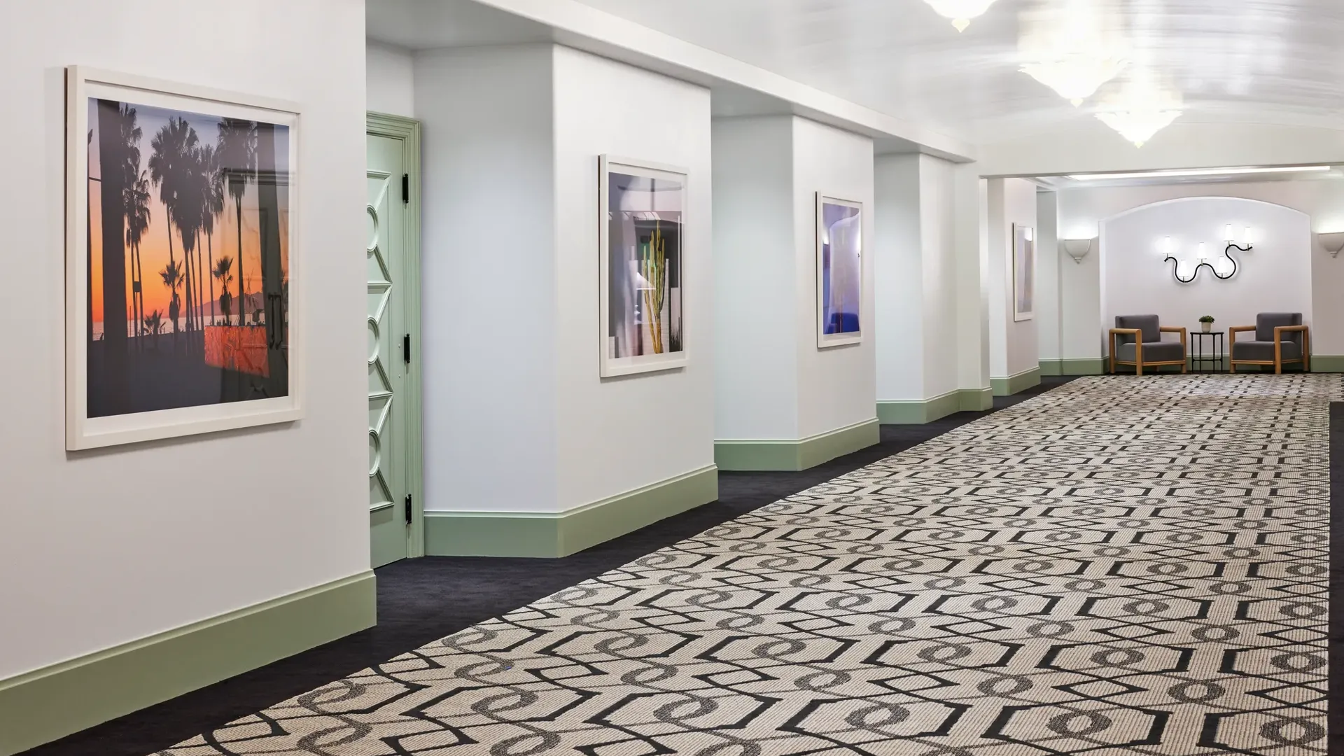 Gallery pre-function corridor with framed art, patterned carpet, mint trim, chandeliers, and small seating nooks.
