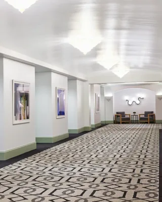 Gallery pre-function corridor with framed art, patterned carpet, mint trim, chandeliers, and small seating nooks.