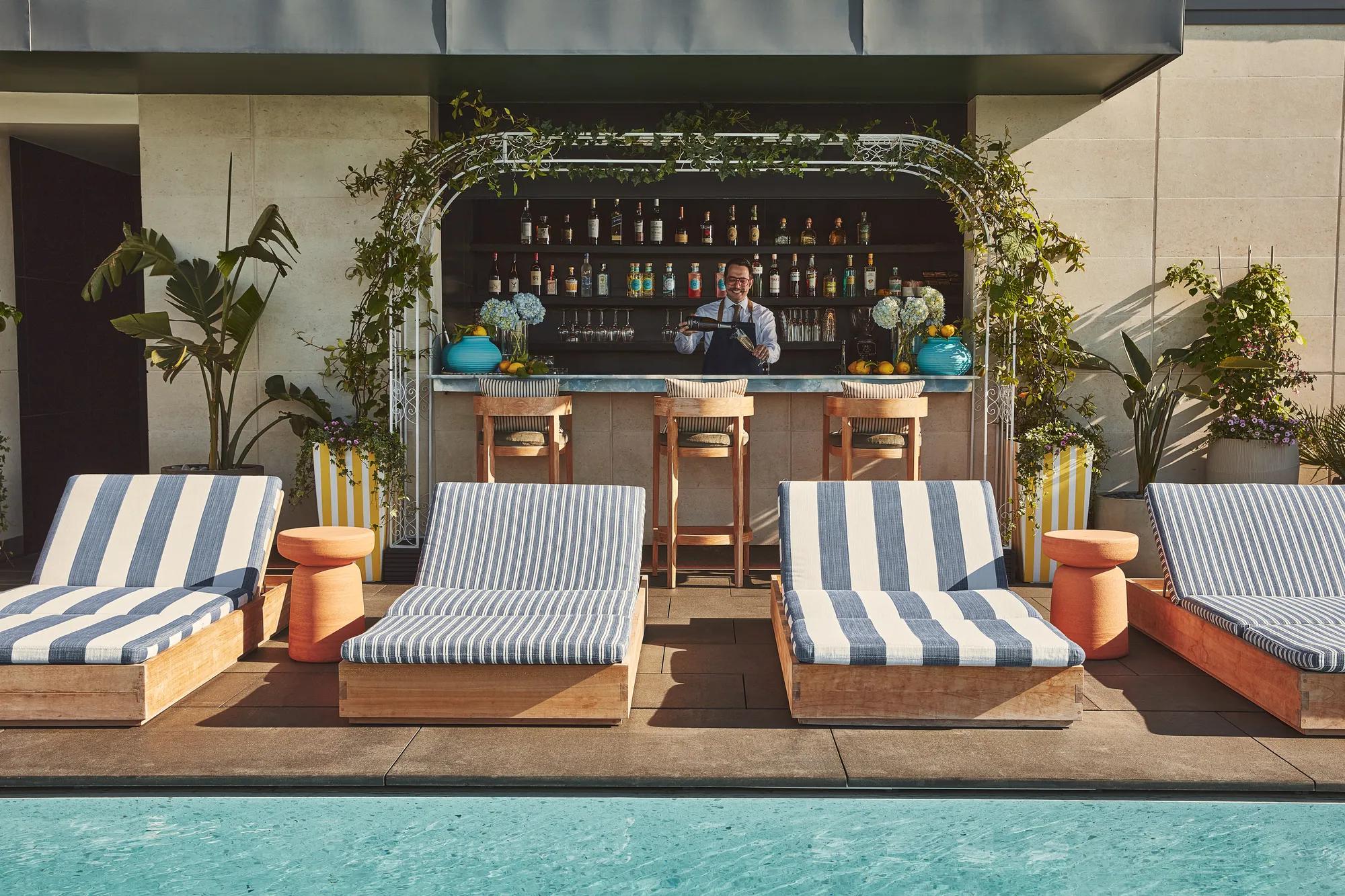 Rooftop pool with striped sunbeds facing a bar framed by greenery, bartender preparing drinks in the background.