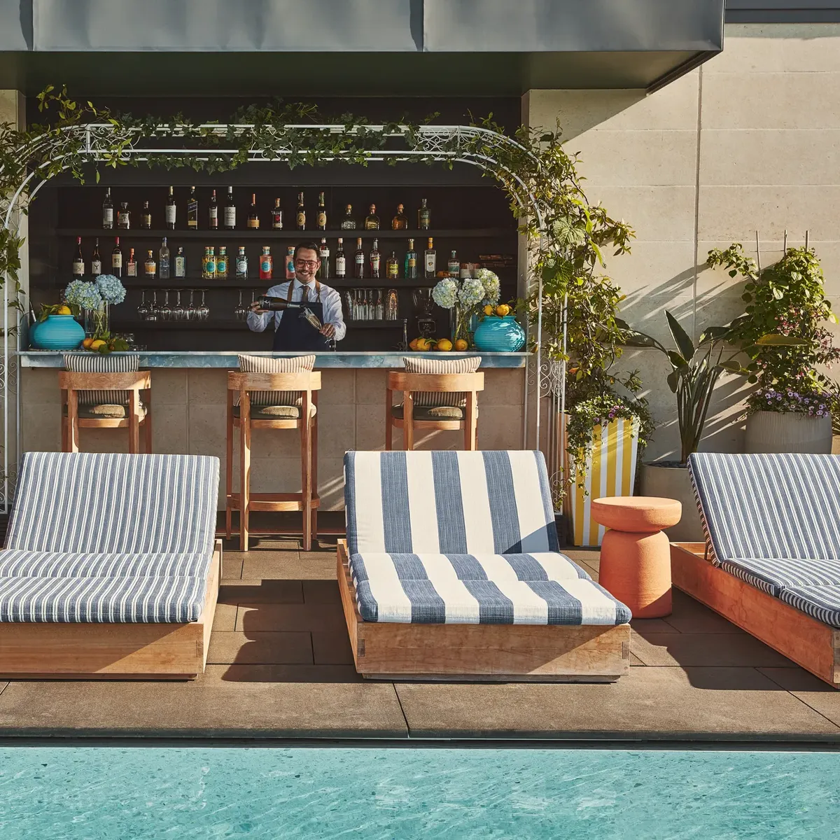 Rooftop pool with striped sunbeds facing a bar framed by greenery, bartender preparing drinks in the background.