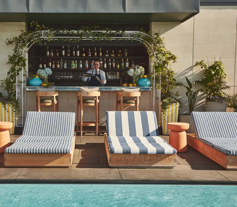 Rooftop pool with striped sunbeds facing a bar framed by greenery, bartender preparing drinks in the background.