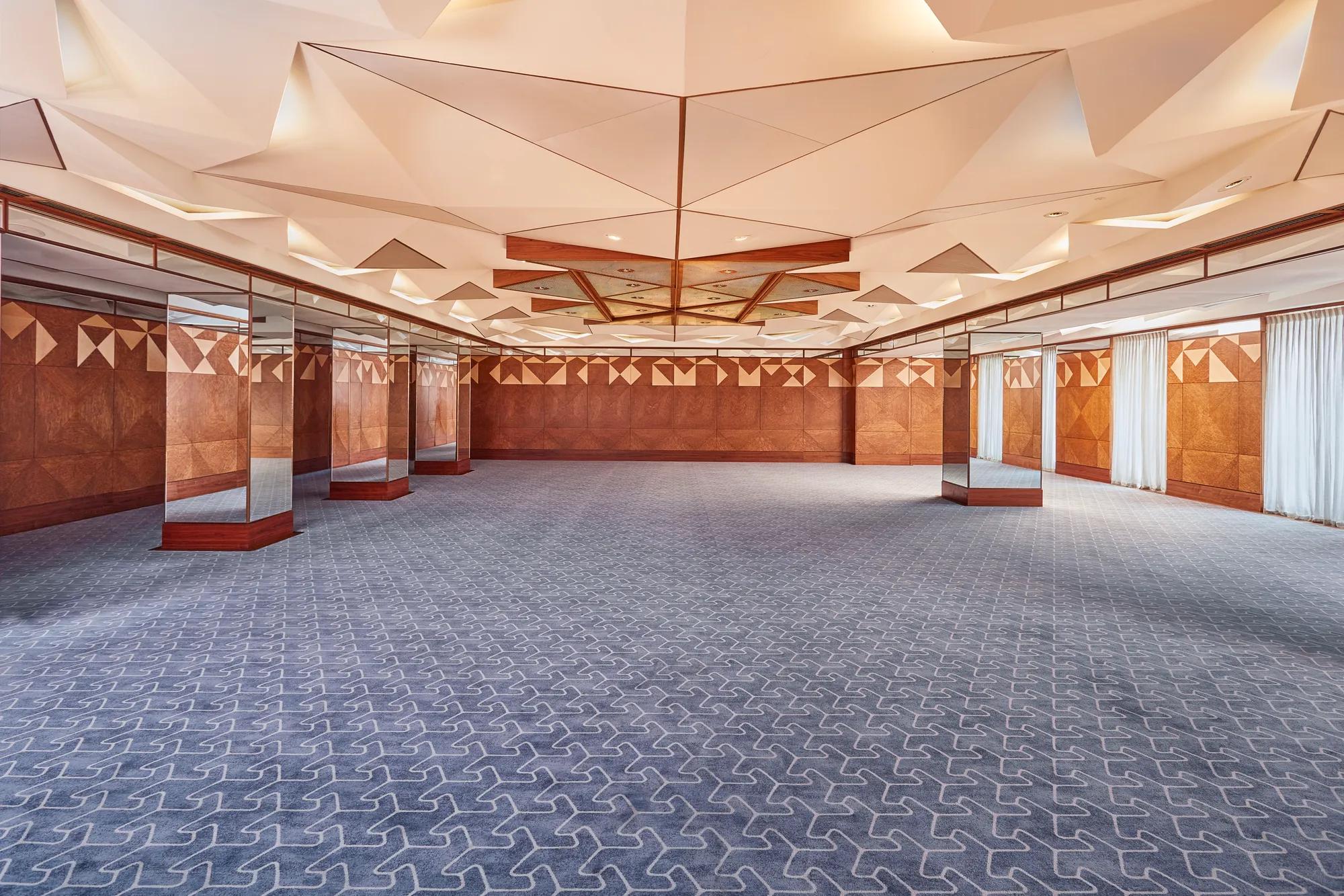 Spacious hotel ballroom with geometric ceiling design, patterned carpet, and mirrored walls for events