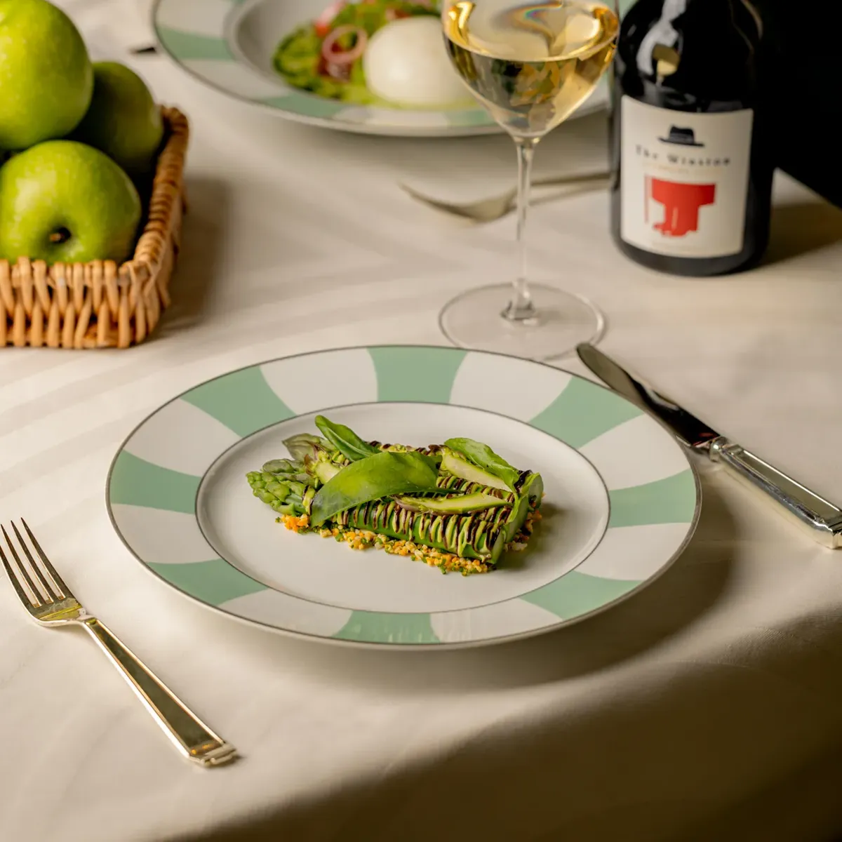 Plated green starter on striped plate with wine glasses, cider bottle, and basket of apples on table.