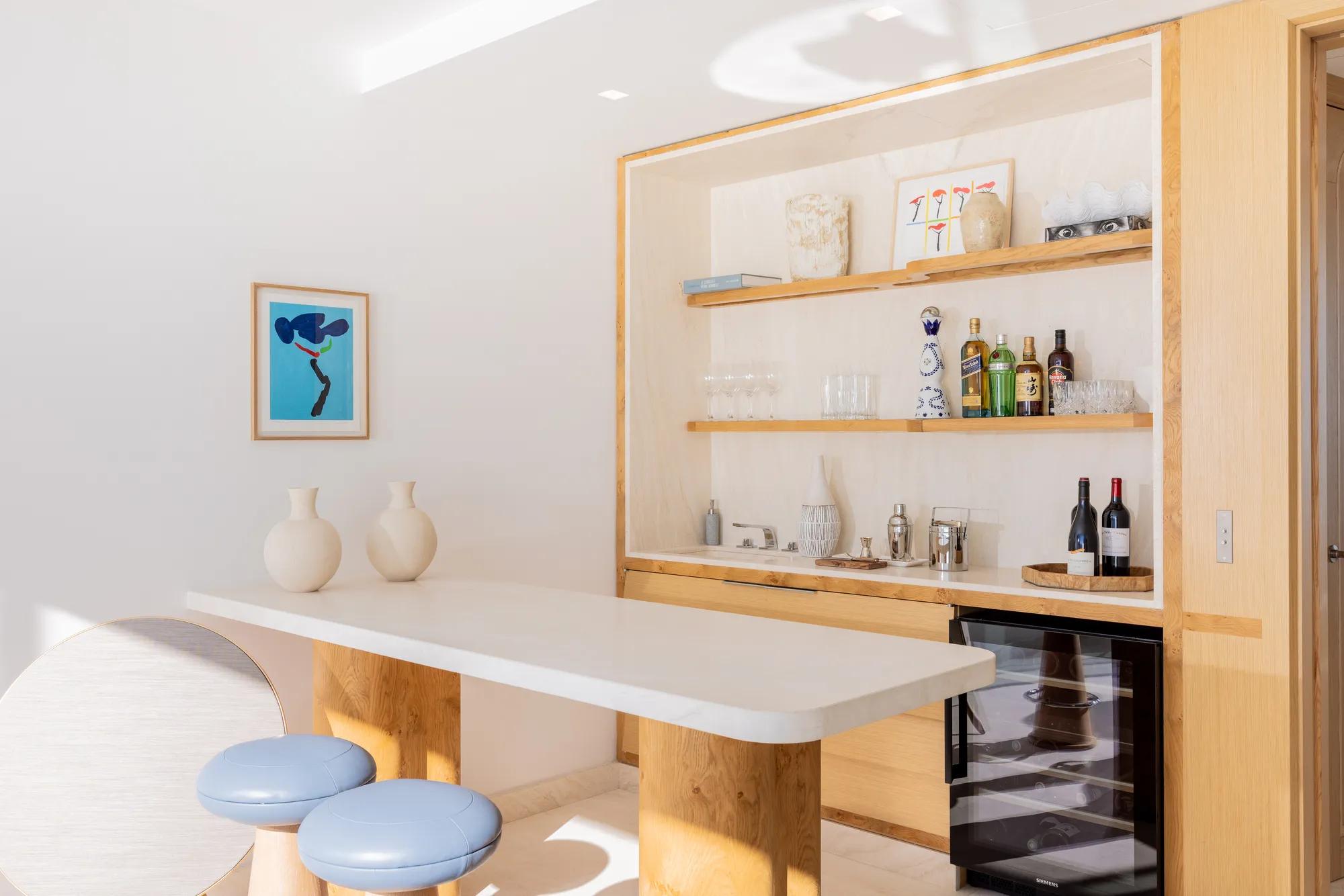 Bar lumineux avec comptoir en marbre, étagères en bois garnies de bouteilles et verres, tabourets bleus et cave à vin en dessous.
