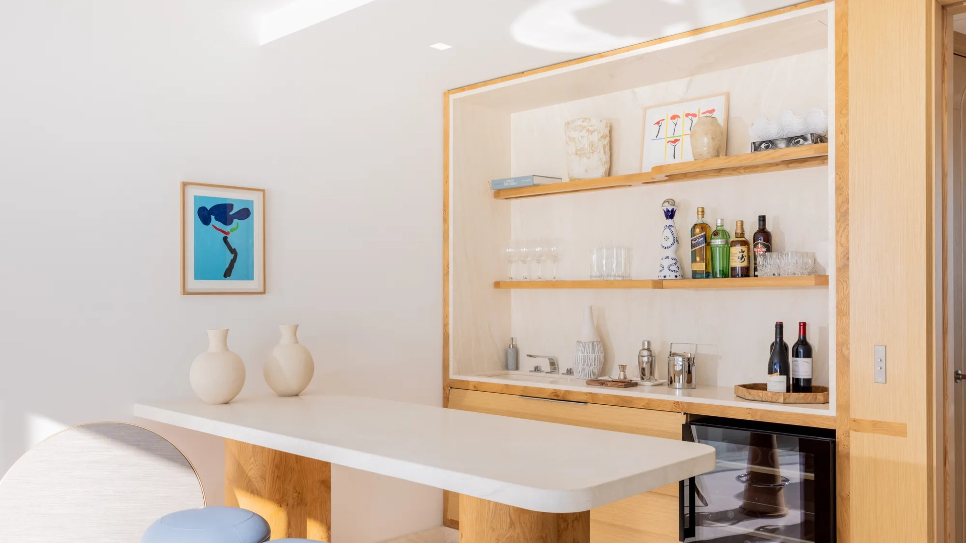 Bar lumineux avec comptoir en marbre, étagères en bois garnies de bouteilles et verres, tabourets bleus et cave à vin en dessous.