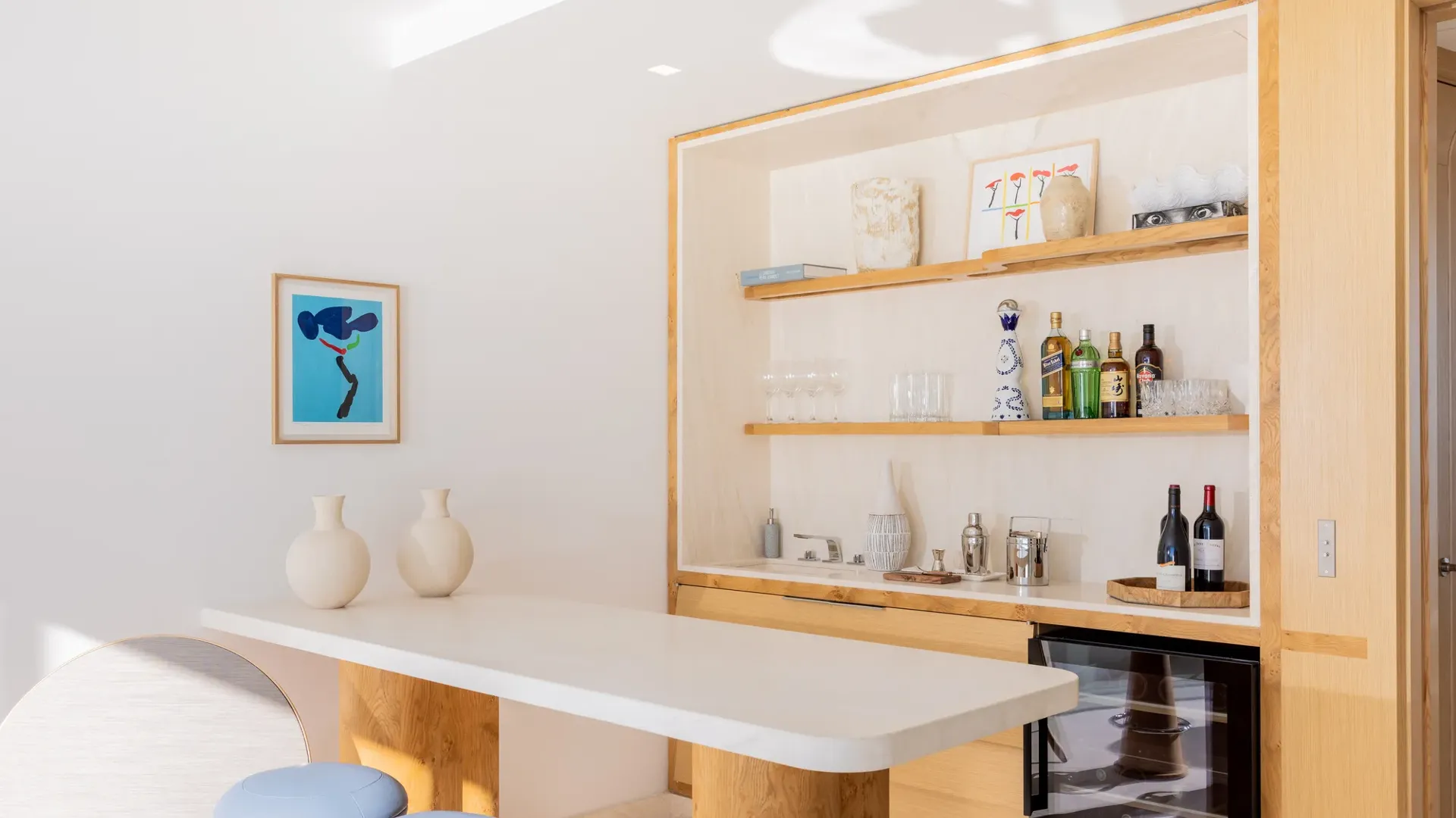 Bar lumineux avec comptoir en marbre, étagères en bois garnies de bouteilles et verres, tabourets bleus et cave à vin en dessous.