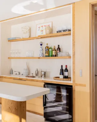 Bar lumineux avec comptoir en marbre, étagères en bois garnies de bouteilles et verres, tabourets bleus et cave à vin en dessous.
