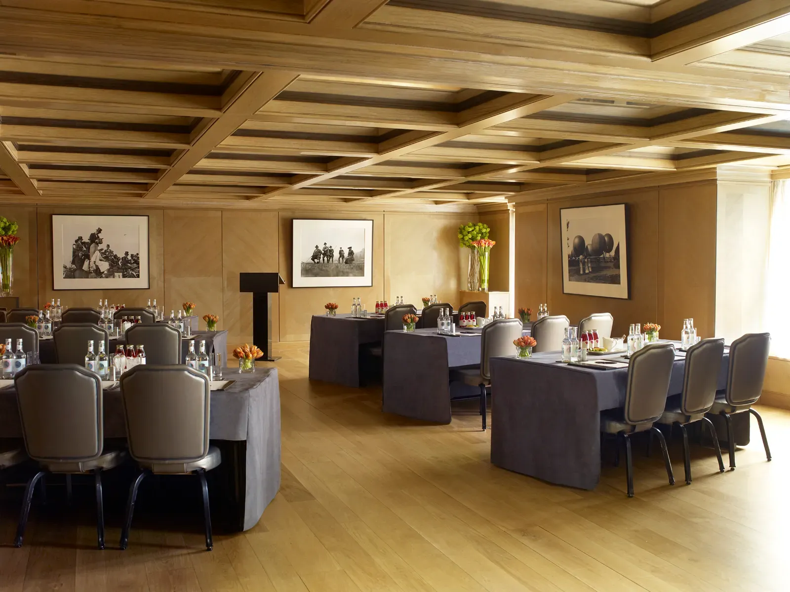 Light-filled meeting room with wood-panelled walls, coffered ceiling, navy-draped tables and bottled water, accented by bright floral arrangements and black-and-white photography.